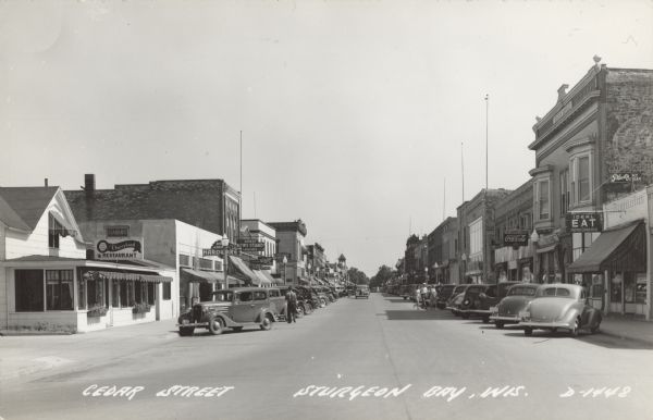 Text on front reads: "Cedar Street, Sturgeon Bay, Wis." Storefronts on a street with automobiles parked diagonally at the curb. Some pedestrians can be seen. Many businesses have signs: "Dairyland Restaurant", "Ideal Eat Home Cooking", "Cabins", "Door County Hardware", "Ahren's News Stand", "Roxana Hotel", "Pleck's Ice Cream", "Western Union" and "Coca-Cola." Cedar Street was renamed 3rd Avenue during WII. Caption reads: Cedar Street Sturgeon Bay, Wis."