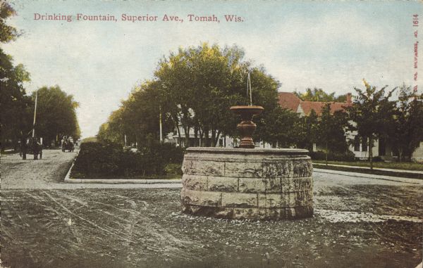 Text on front reads: "Drinking Fountain, Superior Ave., Tomah, Wis." A public drinking fountain on Superior Avenue, a wide unpaved boulevard. Horse-drawn vehicles can be seen in the street and trees, sidewalks and homes fill the neighborhood.