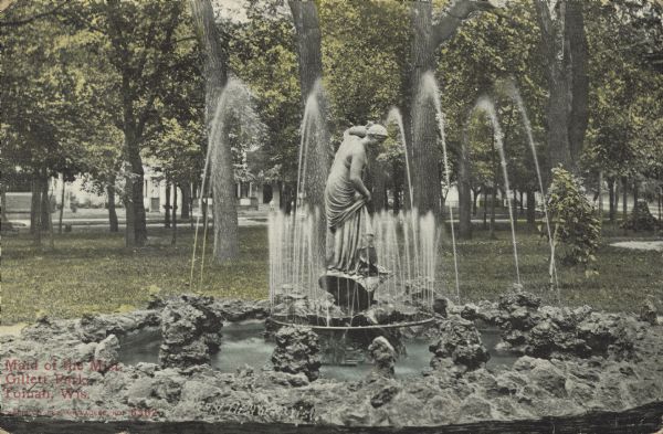 Text on front reads: "Maid of the Mist, Gillett Park, Tomah, Wis." A stone fountain with a statue of a woman in the center, holding a water vessel on her shoulder. In the background is a tree filled park with homes in a neighborhood.