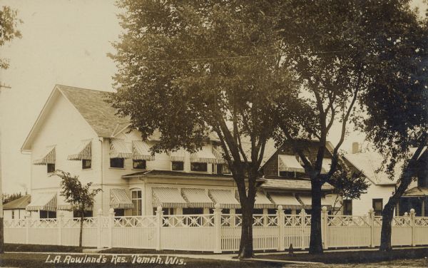 Text on front reads: "L.A. Rowland's Res., Tomah, Wis." A large clapboard lodge with striped awnings and a decorative wooden fence. A sidewalk, lawn and trees are in the foreground.