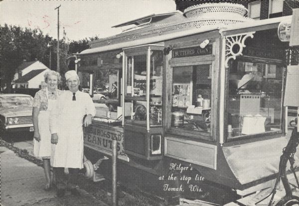 Text on front reads: "Hilger's at the Stoplight, Tomah, Wis." Rubber stamped on the reverse: "Hilger's Pop Corn Wagon. At the Stop & Go Light, Tomah, Wisconsin 54660. Telephone 1-608-372-2975." Ralph and Larue Hilger pose in front of their popcorn truck parked at the curb. Signs on the truck read: "Buttered Corn" and Fresh Roasted Peanuts". Trees and buildings are in the background.