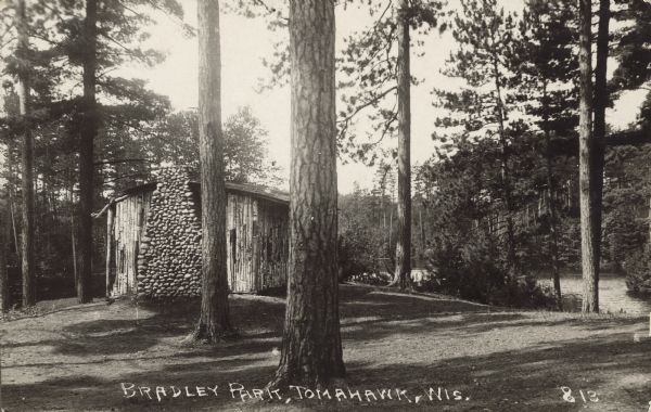 Text on front reads: "Bradley Park, Tomahawk, Wis." The shelter house in Bradley Park with a rubble stone chimney, surrounded by trees. Mirror Lake can be seen in the background.