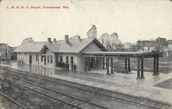 Text on front reads: "C.M. & St. P. Depot, Tomahawk, Wis." The Chicago Milwaukee & St. Paul Railroad station with passengers waiting under the eaves due to rain, the pavement appears to be wet. Tracks are in the foreground and buildings are in the background. 