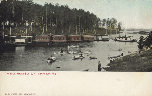 Text on front reads: "Train of House Boats, at Tomahawk, Wis." A train of four houseboats in the water with another boat to pull them. Five rowboats are in the foreground and the opposite shore is a tree covered hill. Tomahawk is located on the Wisconsin River, and is bordered by Lake Mohawksin on the North and West.