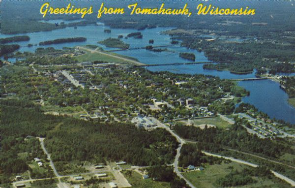 Text on front reads: "Greetings from Tomahawk, Wisconsin." A aerial view showing the city and Lake Mohawksin on the Wisconsin River.