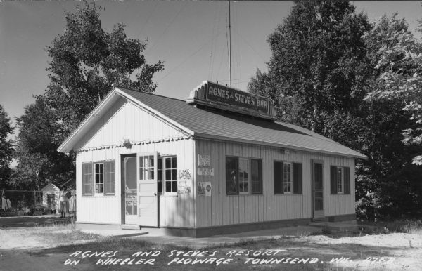 Text on front reads: "Agnes and Steve's Resort on Wheeler Flowage, Townsend, Wis." A bar with a sign on the roof that reads: "Agnes and Steve's Bar". Signs advertising Coca-Cola are at the corner of the building near the door, and hand-lettered signs read: "Please Blow Horn", "Stewing Chicken For Sale", and "Fresh Eggs For Sale". Two women are standing on the left near the corner of the bar, and another woman is walking in front of cabins in the background. Trees surround the resort.