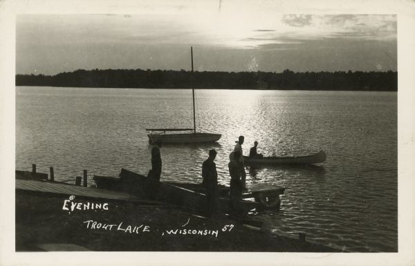 Text on front reads: "Evening, Trout Lake, Wisconsin." As the sun sets, a group of men on a pier watch another man in a canoe on the lake. A sailboat is moored a bit farther out and a rowboat is pulled up on the shore. The far shore is covered with trees. Trout Lake is located between Boulder Junction and Arbor Vitae, Wisconsin.