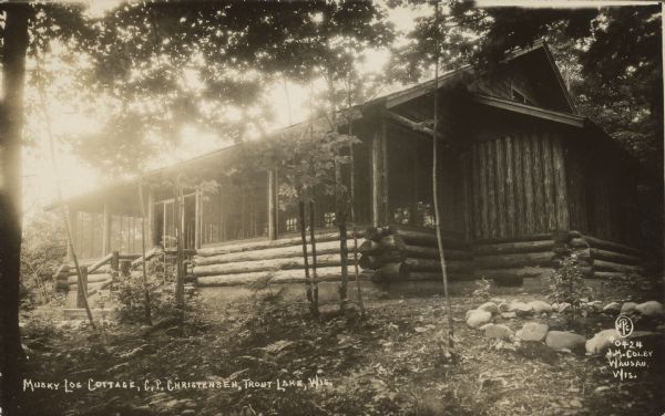 Text on front reads: "Musky Log Cottage, C.P. Christensen, Trout Lake, Wis." A log cabin with a large screened porch, surrounded by trees. Trout Lake is located between Boulder Junction and Arbor Vitae, Wisconsin.