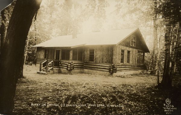 Text on front reads: "Musky Log Cottage, C.P. Christensen, Trout Lake, Wis." A log cabin with large windows and a screened porch. It is surrounded by trees. Trout Lake is located between Boulder Junction and Arbor Vitae, Wisconsin.