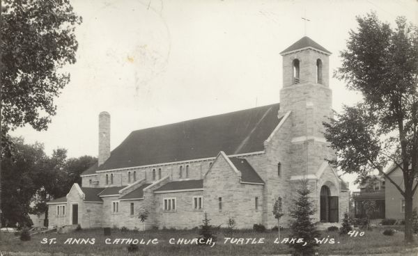 Text on front reads: "St. Anns Catholic Church, Turtle Lake, Wis." A stone church built in 1922, surrounded by trees. More buildings are in the background.