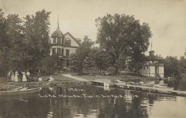 Text on front reads: "Lake House, Twin Lakes, Wis." A group of people are gathered on the lawn at the shore of the lake. Two seated people hold a pennant that reads: "Twin Lakes." There is a long pier with boats tied to it, and an ornate boathouse on the right. In the background is a large dwelling with a tower, sidewalk and trees.