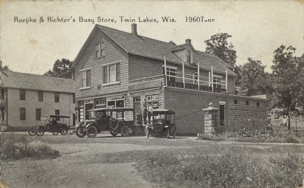 Text on front reads: "Roepke & Richter's Busy Store, Twin Lakes, Wis." Several automobiles are parked in front of the grocery store, and a man is standing on a platform next to a gasoline pump. The store is built of bricks with a dwelling and a deck above. A sign reads: "Meat Market." Another building is on the left.