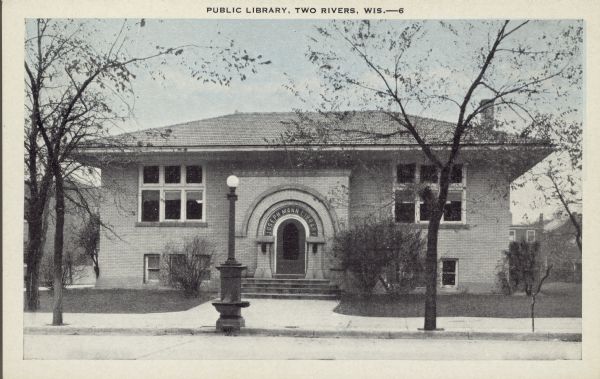 Text on front reads: "Public Library, Two Rivers, Wis." This Carnegie Library was built in 1914 of cream brick in the Prairie School style. It was funded by a $1000 donation from Mrs. Joseph Mann and a $12,500 donation from Andrew Carnegie.