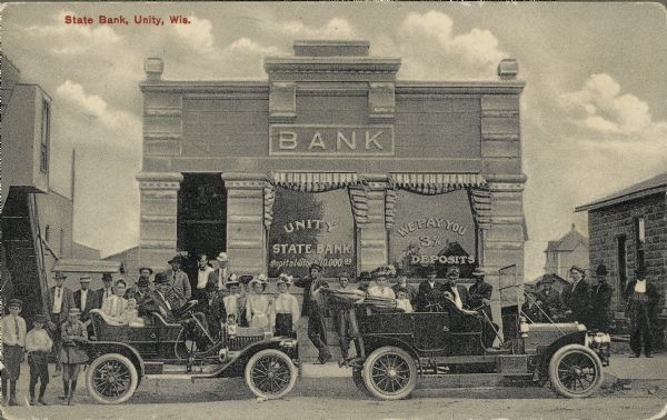 Caption reads: "State Bank. Unity, Wis." A large group of children and adults pose in front of the Unity State Bank, some seated in two automobiles. Signs in the windows read: "Unity State Bank, Capital Stock $10,000.00" and "We Pay You 3% On Time Deposits."