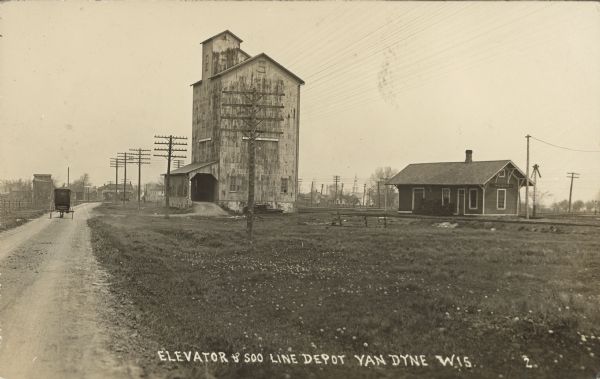 Text on front reads: "Elevator & Soo Line Depot, Van Dyne, Wis." A horse-drawn vehicle is on an unpaved road beside the wooden grain elevator and train depot. A sign on the elevator reads: "Manitowoc - Malting - Co." Buildings of the town are in the distance.