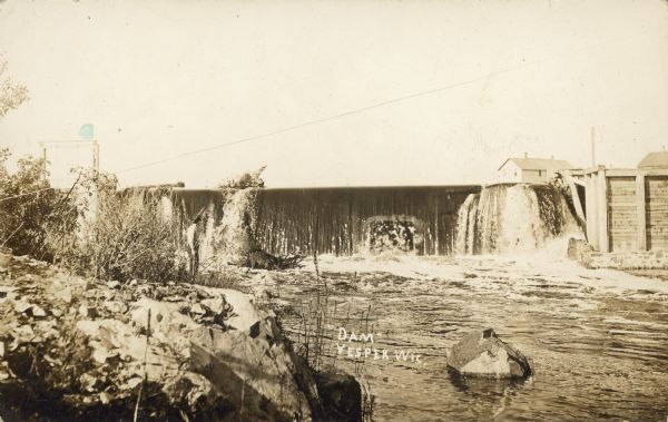 Text on front reads: "Dam, Vesper, Wis." The privately owned dam was built on the Hemlock Creek River in 1944 for recreational purposes. Buildings are beyond the dam on the horizon.
