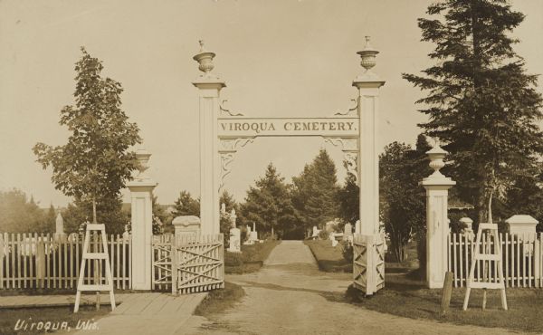 Text on front reads: "Viroqua, Wis." The driveway gate sign reads: "Viroqua Cemetery." The gate, sign and fence are painted a light color. There are many trees.