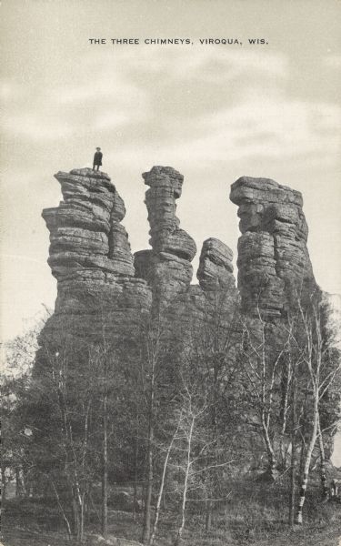 Text on front reads: "The Three Chimneys, Viroqua, Wis." A man stands on top of one of the pillars in the Three Chimneys rock formation, also known as Chimney Rock. The base is surrounded by trees.