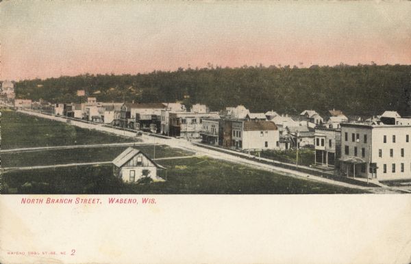 Text on front reads: "North Branch Street, Wabeno, Wis." Elevated view of a street and sidewalk with most of the storefronts and buildings on the far side. A hill with trees fills the horizon.
