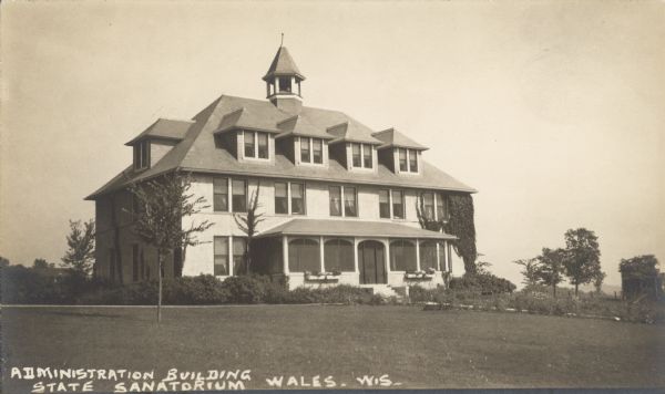 Text on front reads: "Administration Building State Sanatorium, Wales, Wis." A three-story building with dormers and a porch, surrounded by shrubs and trees. The Wisconsin State Tuberculosis Sanatorium was also known as “Statesan." It was the only state run Sanatorium in Wisconsin. It opened on November 7, 1907 and closed in the fall of 1957.