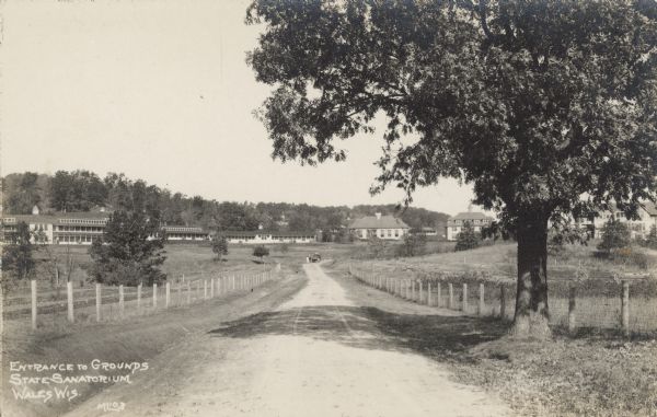 Text on front reads: "Entrance to Grounds, State Sanatorium, Wales, Wis." A view down the road towards the Wisconsin State Tuberculosis Sanatorium, also known as “Statesan", with multiple buildings along the base of tree-covered hills. Fences line the road and a large tree is in the right foreground. It was the only state run Sanatorium in Wisconsin. It opened on November 7, 1907 and closed in the fall of 1957.