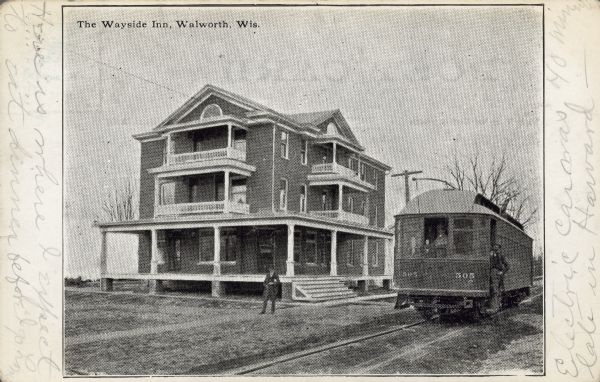 Text on front reads: "The Wayside Inn. Walworth, Wis." Also known as the Wayside Hotel, it was built in 1902 of brick with porches on all three levels. A man is standing in front of the building. A passenger car and  railroad tracks are on the right, with a man inside the window and another standing on the step outside the doorway.