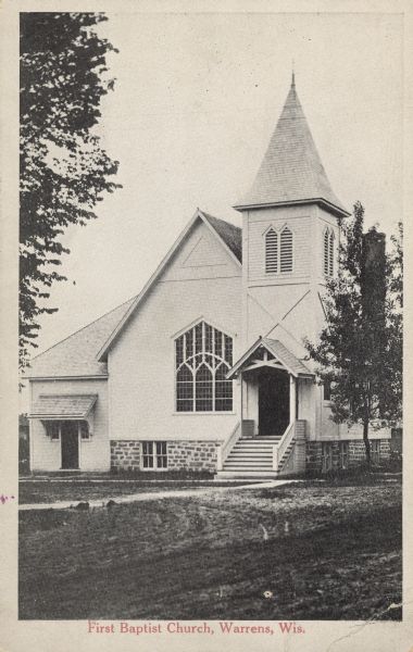 Text on front reads: "First Baptist Church, Warrens, Wis." A clapboard church built in the Queen Anne style with ornate stained glass window, belfry and trees.