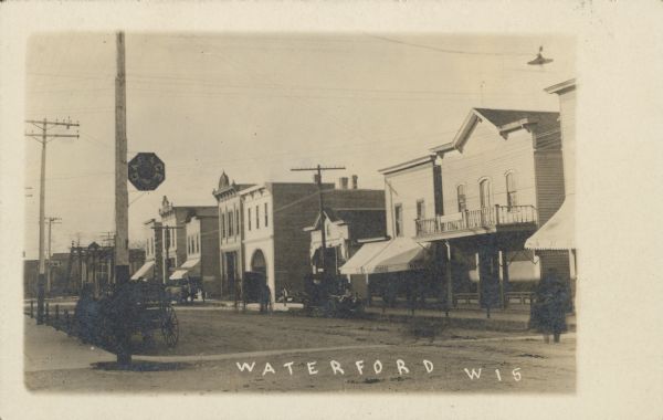 Text on front reads: "Waterford, Wis." Storefronts along a street, with automobiles and horse-drawn vehicles parked at the curb. Pedestrians are on the street and sidewalks. A sign that reads: "Garage" is mounted on a utility pole on the left.