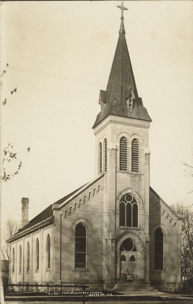 Text on front reads: "German Lutheran Church". Two young girls are sitting on the top of the steps at the front door. Also known as "St. John's Evangelical Lutheran Church." The cream brick church was built in 1892 in the Gothic Revival style. In 1982 it was demolished and replaced by a new church.