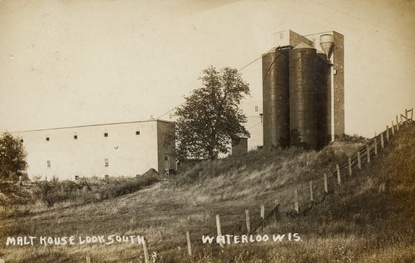 Text on front reads: "Malt House Look South. Waterloo, Wis." Three buildings with two large silos, this malt house was originally constructed in 1901. The silos are on a hill above two of the buildings with a fence on the right.