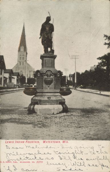 Text on front reads: "Lewis Indian Fountain, Watertown, Wis." The fountain stands on a brick street with buildings, a church and trees in the background. The cast zinc statue of an unknown Native American Chief, manufactured by J.L. Mott, was gifted to the city by Robert E. and Fanny Lewis in memory of their son, Clifton, in 1896. It stood on top of an ornamental drinking fountain. In 1925 in was struck by an automobile and damaged beyond repair. An exact duplicate was ordered from the original manufacturer and placed in Union Park. In the 1960s, the city decided to remove the statue and it was given to the Watertown Historical Society. The Society placed it near the Octagon House in 1964 where it remains today.