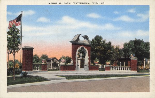 Text on front reads: "Memorial Park, Watertown, Wis." A memorial park with a brick and stone arch, balustrades and columns. There are planters with flowers, an American flag and trees.