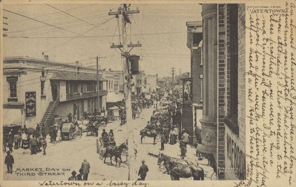 Text on front reads: "Market Day on Third Street. Greetings from Watertown, Wis." Elevated view of a busy street scene on market day, with many pedestrians, horses, horse-drawn buggies and wagons. On the left is "Deutsches Dorf" saloon.