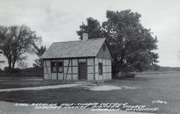 Text on front reads: "Karl Zettler Half-Timber Cottage, Ozaukee County Pioneer Village, Waubeka, Wisconsin." Built in 1849, the cottage was moved to the Ozaukee County Pioneer Village from the original location west of Wabeka in the 1960s.