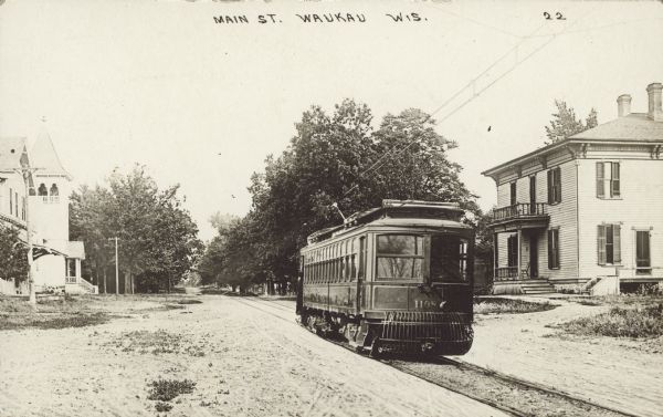 Text on front reads: "Main Street, Waukau, Wis." An unpaved street with a street car and tracks running down the center. A church is on the left and a home is on the right.