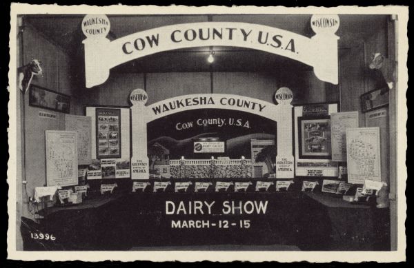 Text on reverse reads: "We invite you to attend Waukesha County Dairy Show. March 12-15, 1940. Sales Pavilion, Waukesha, Wis. Judging days, March 13-14. For live stock breeders, judging of Holsteins, Jersey and Brown Swiss on 13th. Judging of Guernsey Ayrshire on 14th. Programs 4 days and 4 nights." A display advertising a Dairy Show with many maps, posters and cutout signs. Some of the text reads: "Cow Country U.S.A.", "Waukesha County", "The Holstein Capital of America" and "The Guernsey Capital of America." The postcard has a deckled edge.