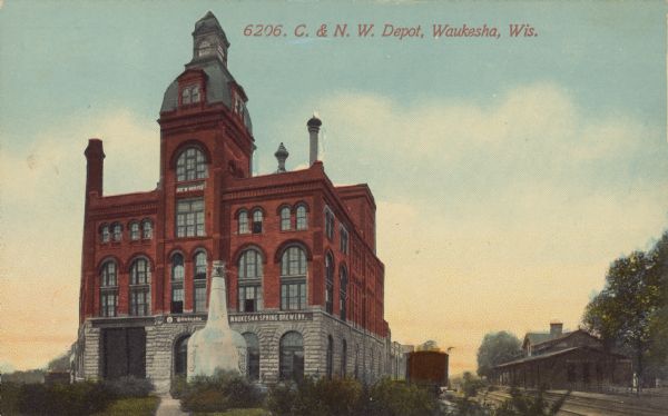 Text on front reads: "C. & N.W. Depot, Waukesha, Wis." The Waukesha Spring Brewery building is on the left and the Chicago & North Western Depot is on the right. There is an oversized beer bottle in front of the building near the sidewalk, which appears to be advertising Fox Head Beer. The brewery was established in 1893 and changed owners and names many times. Some names include: "Waukesha Spring Brewing Company", "Imperial Spring Brewery", "Waukesha Imperial Spring Brewing Company", Milwaukee-Waukesha Brewing Company" and Fox Head Spring Beverage Company."
