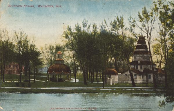 Text on front reads: "Bethesda Spring, Waukesha, Wis." Bethesda Spring is located inside an ornate pavilion surrounded by trees. The Terrace Hotel can be seen on the left and another building on the right. The word "Bethesda," seen on the left, is on the bank of a pond. In the foreground is the Fox River. Between 1868 and 1918, 60 mineral springs were located here. People would travel here to "enjoy the waters."