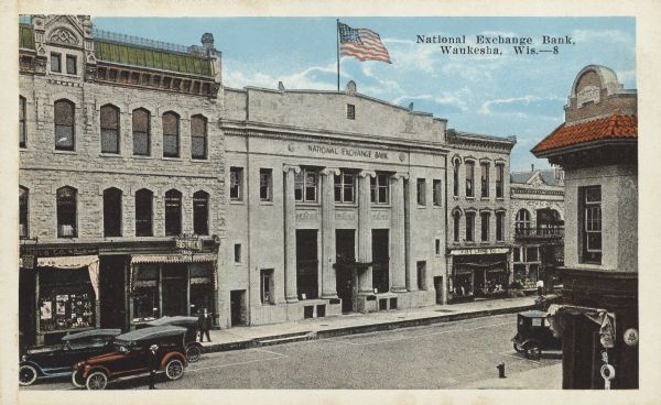 Text on front reads: "National Exchange Bank, Waukesha, Wis." Built in 1921 in the Neoclassical style. Located on West Main Street between other brick and stone buildings. Pedestrians are on the sidewalks and automobiles on the street.