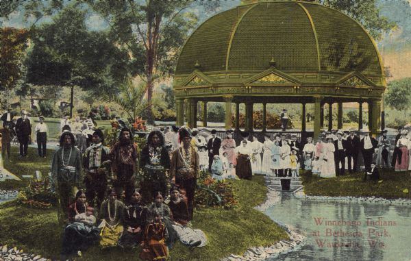 Text on front reads: "Winnebago Indians at Bethesda Park, Waukesha, Wis." A group of Ho-Chunk pose at Bethesda Park as a crowd of tourists looks on. There are five men, five women, one youth and two infants. They are wearing indigenous clothing. Between 1868 and 1918, 60 mineral springs were located here. People would travel here to "enjoy the waters."