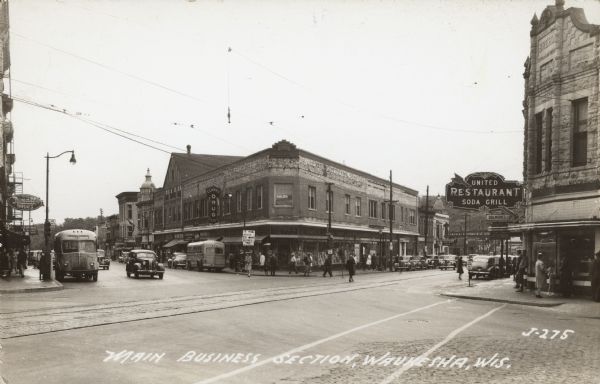 Text on front reads: "Main Business Section, Waukesha, Wis." A busy street scene with pedestrians, automobiles and buses. A railroad track runs down the center of the street. Many businesses can be seen. Several signs read: "United Restaurant Soda Grill", "Delmores Ladies Shop", "Drugs for Less", "Salen", "Clarke Drug Co.", "Rexall Drugs" and "Loans."