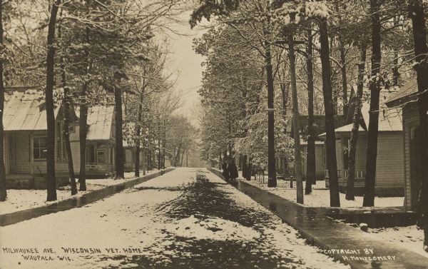 Text on front reads: "Milwaukee Ave. Wisconsin Vet. Home. Waupaca, Wis." Two women walk on a sidewalk along a snow-covered, unpaved street. On each side are cottages surrounded by trees, built for injured Civil War Veterans and their wives. The facility began operations in 1888. Today the Veterans Cottages Historic District is listed on the National and State Register of Historic Places.