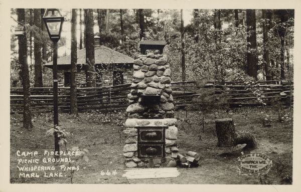 Text on front reads: "Camp Fireplace - Picnic Grounds - Whispering Pines - Marl Lake." A seal at lower right reads: "Chain o' Lakes Waupaca, Wis. The Killarneys of America. Estberg Photo Service." A camp fireplace with a lamppost on the left and a log seat on the right. In the background is a split rail fence, stone building and trees.