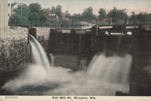 Text on front reads: "Fall Mill St., Waupaca, Wis." Water flowing over a dam on the Waupaca River. There are advertising signs on the dam and  buildings can be seen on the far shoreline.