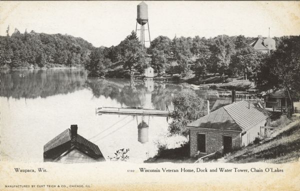 Text on front reads: "Waupaca, Wis. Wisconsin Veteran Home, Dock and Water Tower, Chain O'Lakes." Elevated view from hill of a man on a pier, walking towards cottages at the Veterans Home. A water tower is across Rainbow Lake. The Wisconsin Veterans Home was built for injured Civil War Veterans and their wives. The facility began operations in 1888. Today the Veterans Cottages Historic District is listed on the National and State Register of Historic Places.