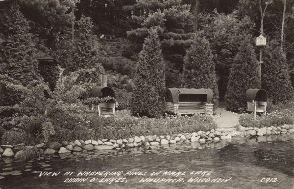 Text on front reads: "View at Whispering Pines on Marl Lake, Chain O' Lakes, Waupaca, Wisconsin." Outdoor seating with canopies and a birdhouse on the shore of Marl Lake. The shoreline is lined with rocks and a flower garden. A paved path winds down to the water. A hill landscaped with foliage, shrubs and trees is in the background. The corner of a roof can be seen on the left.