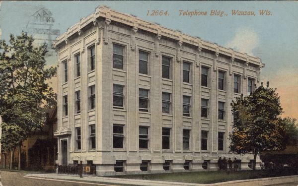 Text on front reads: "Telephone Bldg., Wausau, Wis." A stone building with a decorative parapet has three stories plus a lower level. Four men are standing against the wall on the right and another man is standing at the left corner. There are trees on both sides. A sign in an upper window reads: "Telephone Building."