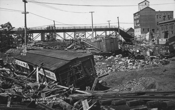 Text on front reads: "Below the High Bridge After the Flood. Wausau, Wisconsin." Railroad cars were swept away when dams at Brokaw and Merrill failed on the Wisconsin River, sending a wave of water through Wausau. Many bridges were damaged. Several men and a boy can be seen observing the piles of timbers, rocks, rail cars and pools of water. On the right is a flour mill with the signs: "Northern Milling Company" and "Use Pure Quality Flour All Ways."