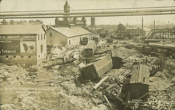Text on front reads: "Below the High Bridge After the Flood. Wausau, Wisconsin." Railroad cars were swept away when dams at Brokaw and Merrill failed on the Wisconsin River, sending a wave of water through Wausau. Many bridges were damaged. Many people can be seen observing the piles of timbers, rocks, rail cars and pools of water. On the left is a building with "Grocery Company" and a bull painted on it. In the background is the old, red brick, Victorian style City Hall with turrets and a clock tower. Built in 1885, it was demolished in 1966.