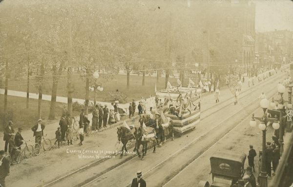 Text on front reads: "Granite Cutters in Labor Day Parade, Wausau, Wisconsin." Elevated view of a parade on a city street with street car tracks, near an urban park area. A float pulled by four horses is followed by members of the G.C.I.A., Granite Cutters International Association. Spectators, bicycles, and automobiles line the streets.